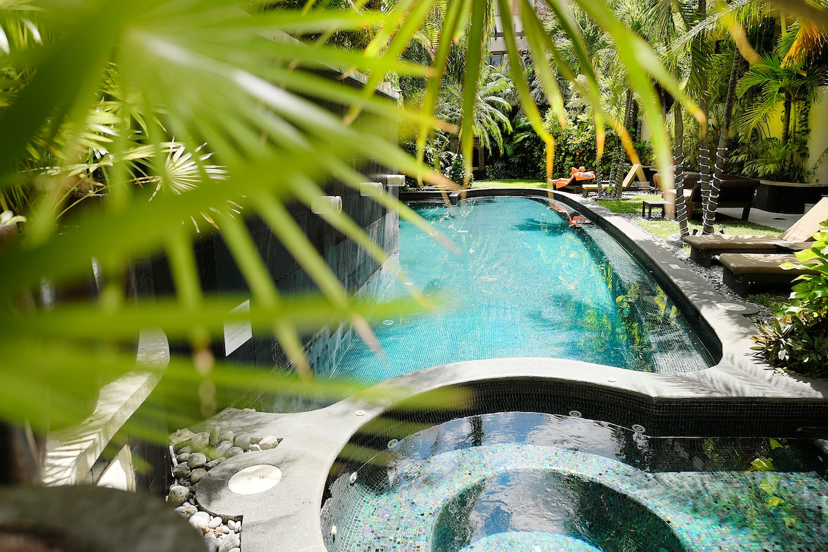 A tranquil swimming pool is framed by lush tropical plants, creating a serene atmosphere. The pool features a mosaic-tiled design with curved edges and is surrounded by comfortable loungers and vibrant greenery, inviting relaxation in a peaceful setting.