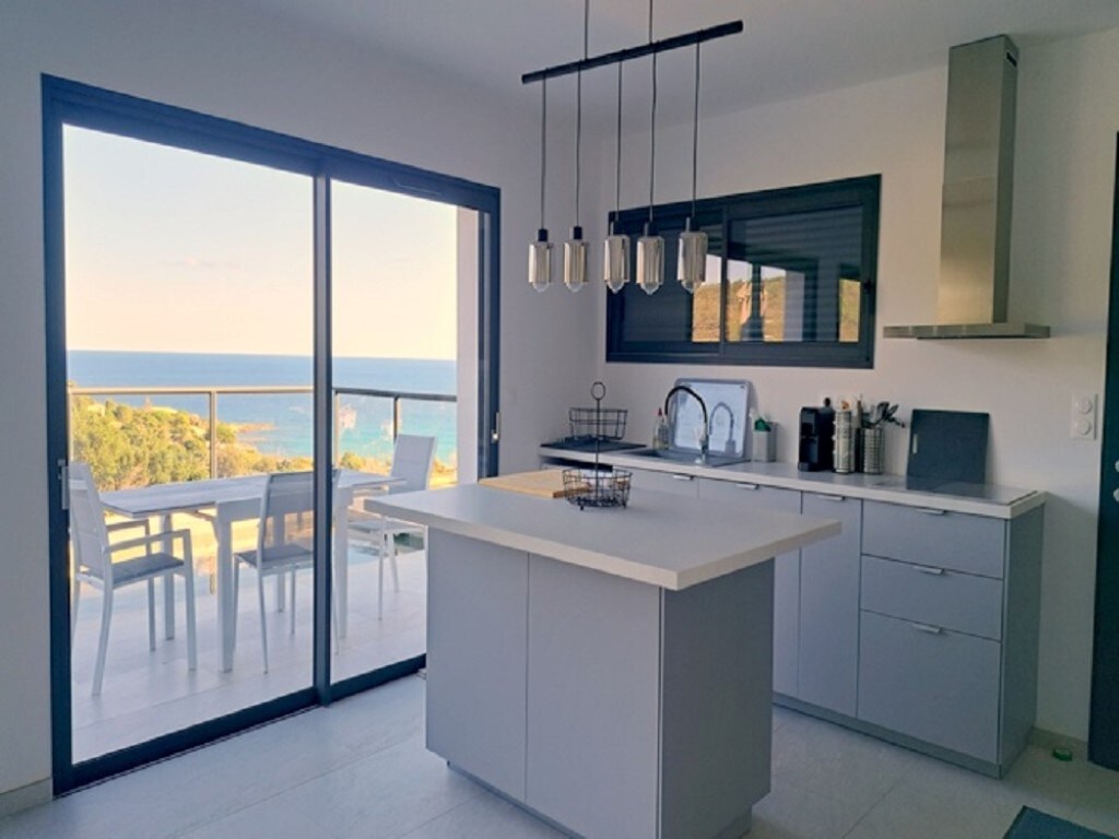 The kitchen features a central island and modern appliances, complemented by large sliding glass doors that open to a terrace with a view of the sea. Natural light floods the space, enhancing the sleek design and functionality.
