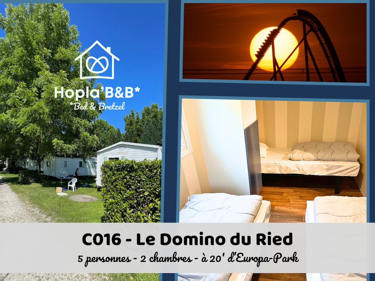 A mobile home is set within a green landscape, with a pathway leading to it. Two rooms are visible; one with a double bed and another featuring three twin beds, suitable for guests. The exterior shows a blend of natural light and trees.