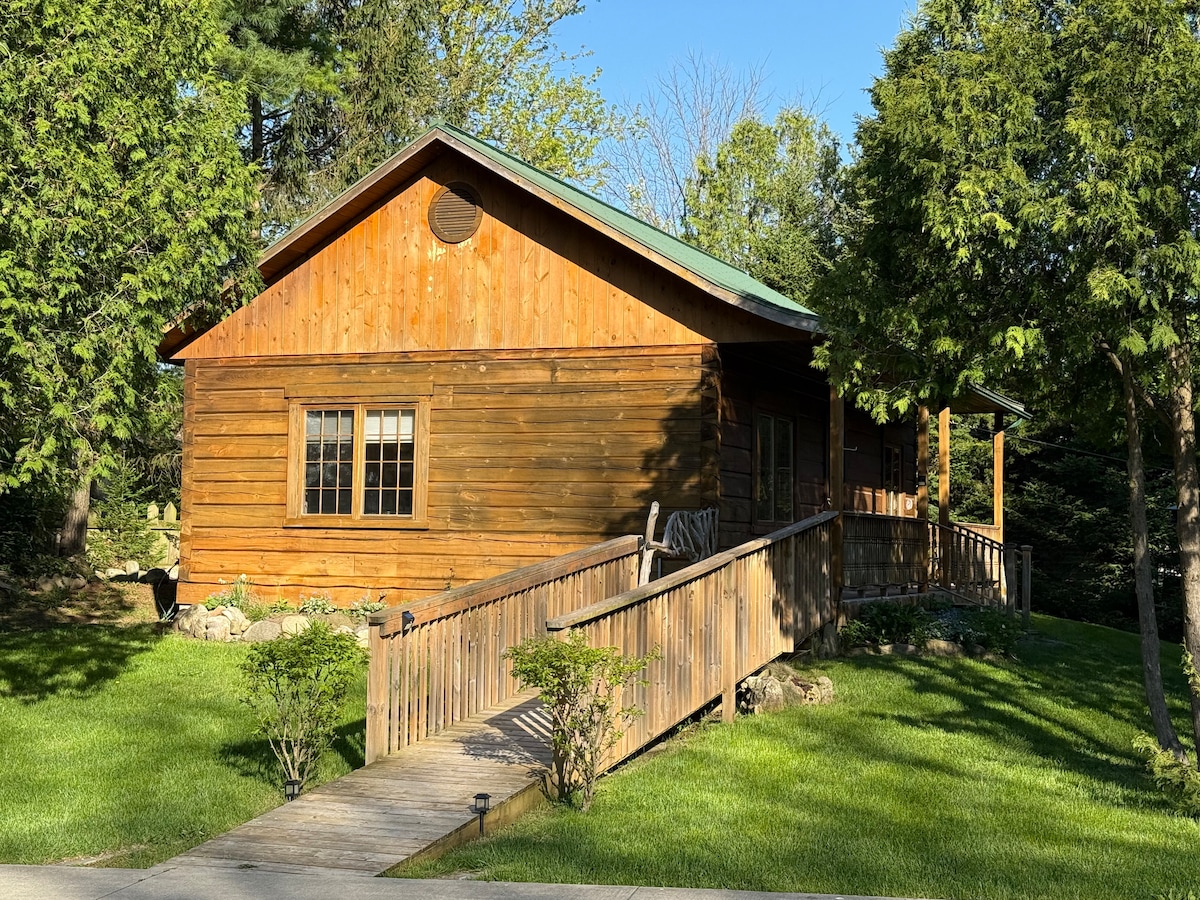 A rustic wooden cabin is framed by lush greenery and mature trees. A wooden walkway leads to the covered porch, where seating is provided. Large windows allow natural light to enter, enhancing the warm appearance of the structure. The surrounding lawn is well-maintained.