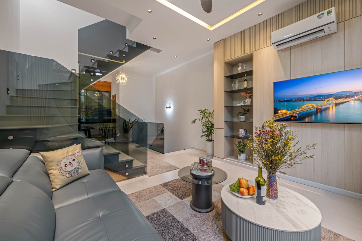 A modern living space features a comfortable, grey sofa with a decorative pillow. A sleek coffee table with fresh fruits and a bottle is positioned in front. Expansive glass stairs lead to the upper levels, and a large television displays vibrant images of the cityscape.