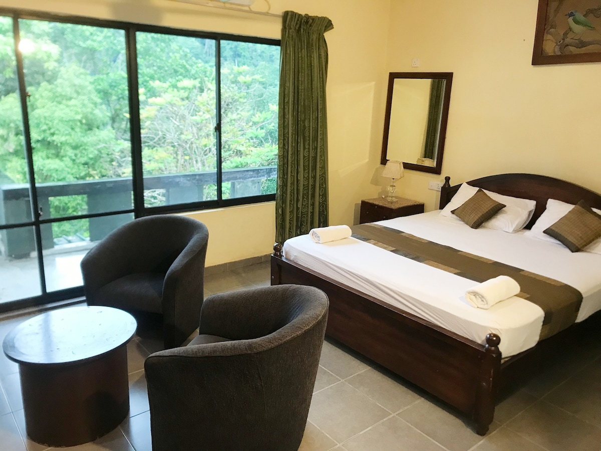 A spacious room features a large bed flanked by two rolled towels. Two comfortable armchairs are positioned around a small round table. A large window offers views of the lush greenery outside, with soft natural light illuminating the space.