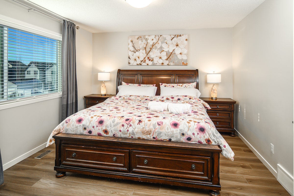 A spacious bedroom features a large bed with a floral comforter and elegant wooden furniture. Two bedside lamps provide warm lighting. A window with light curtains allows natural light to fill the room, while decorative wall art adds a touch of style.