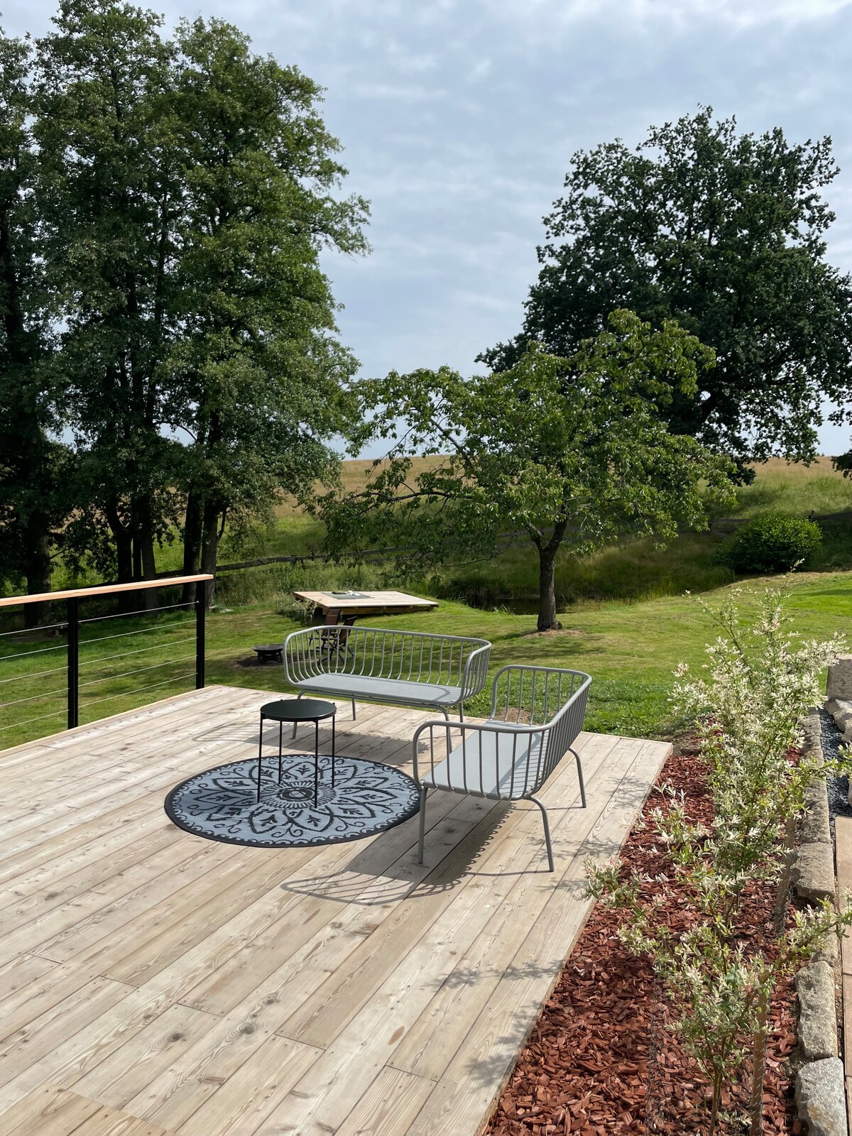An outdoor terrace is shown, featuring a wooden deck with two modern chairs and a small table. A patterned rug lies beneath the table. Lush greenery, including trees and a grassy area, is visible in the background, enhancing the serene atmosphere.
