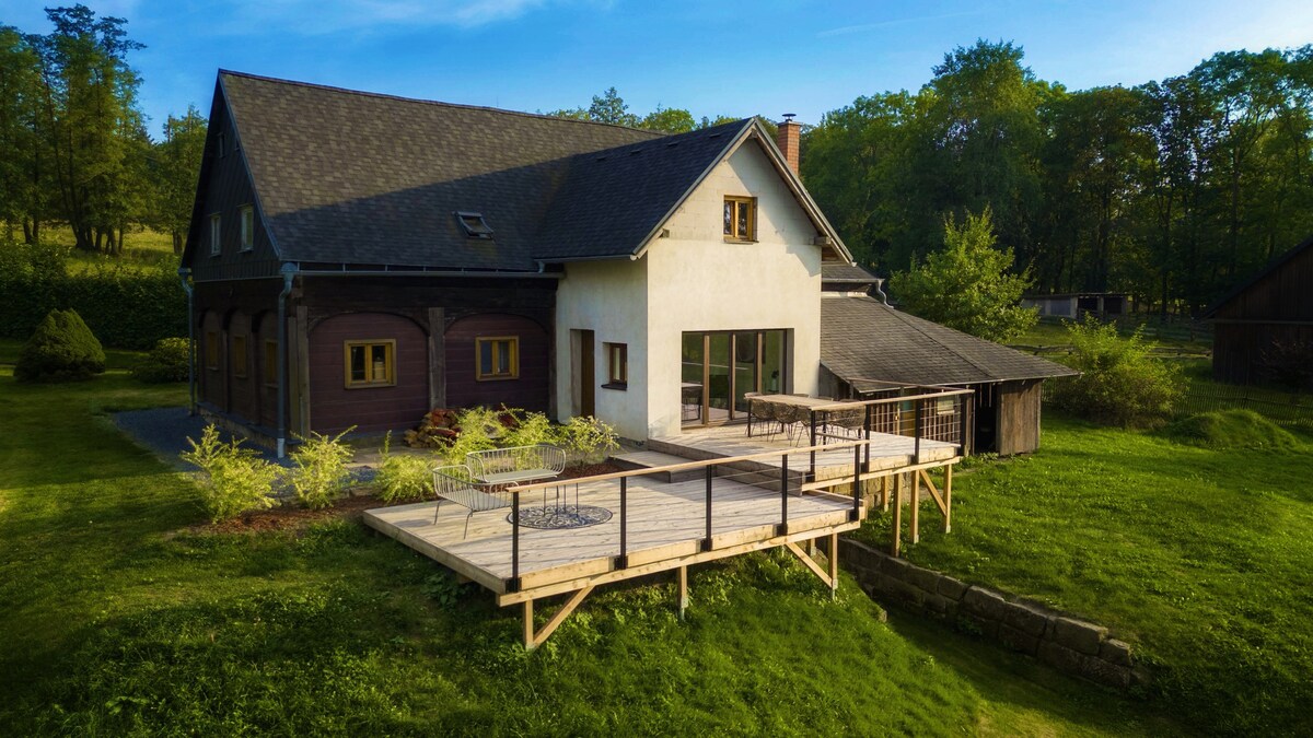 The exterior of an old farmhouse is captured in natural light, featuring a large wooden deck that extends from a modern entrance. Lush greenery surrounds the property, enhancing the sense of privacy and tranquility. The structure combines traditional and contemporary architectural elements.