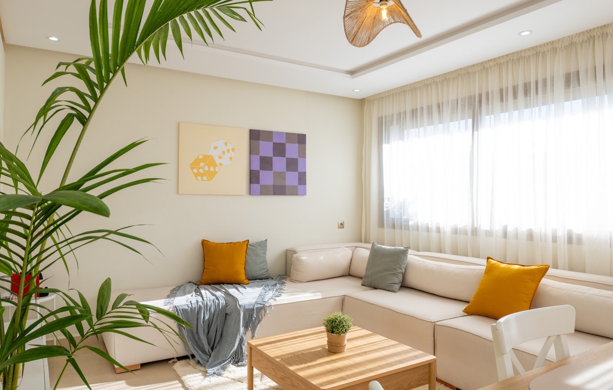 A bright seating area features a modern, L-shaped couch adorned with two orange cushions. A minimalistic wooden coffee table rests on a light rug, complemented by a small potted plant. Natural light pours in through large windows, enhanced by sheer curtains.