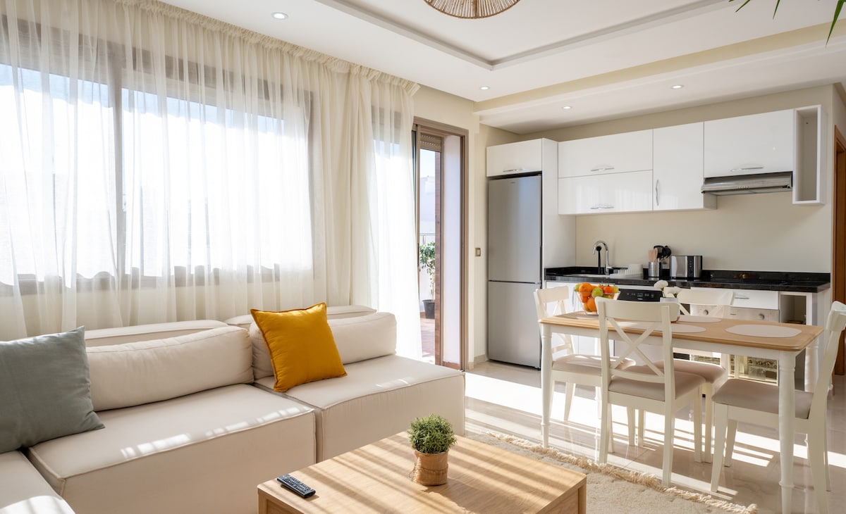 A bright living area features a light-colored sofa accented by pillows and a wooden coffee table. Natural light floods in through large windows adorned with sheer curtains. The adjacent dining area includes a simple table and chairs, with a kitchenette visible in the background.