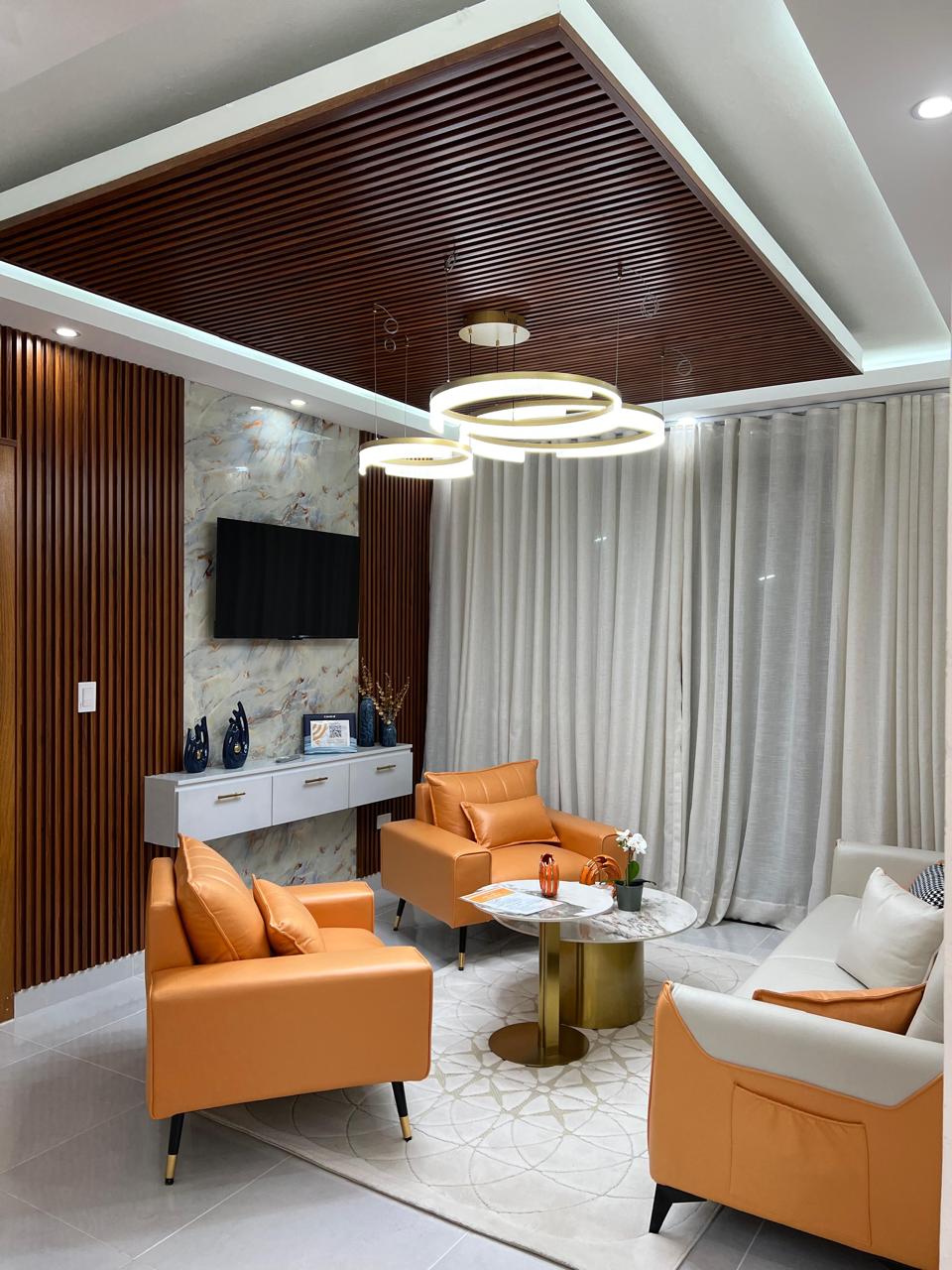 A modern living area features two orange armchairs and a light-colored sofa. A round coffee table occupies the center, surrounded by neutral-toned curtains. A flat-screen TV is mounted on the wall, while decorative accents are displayed on an adjacent sideboard.