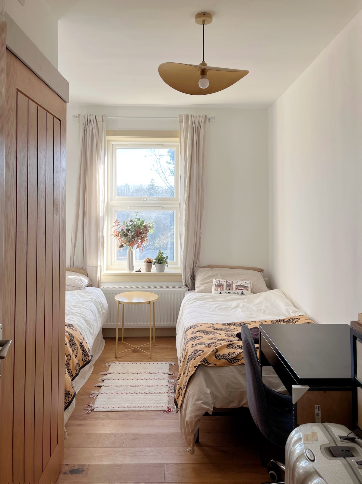 A twin bedroom is shown with two beds featuring light bed linens. A small round table stands between the beds, complemented by a window that allows natural light to fill the room. Simple décor and a warm wooden floor create a welcoming environment.