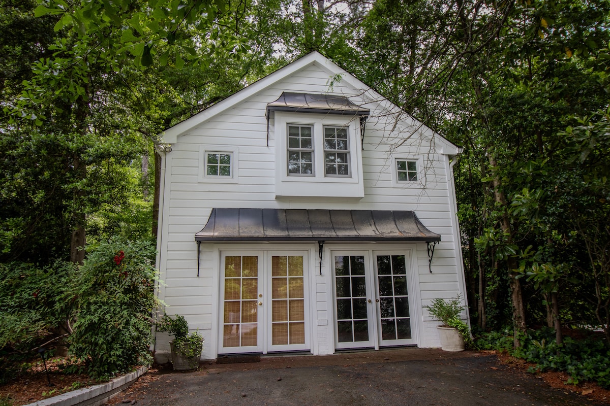 A charming two-story white house is surrounded by lush greenery. Large windows provide natural light and visibility, while the entrance features a double door framed by elegant landscaping. The sloped roof and small awning complement the overall aesthetic of the home.