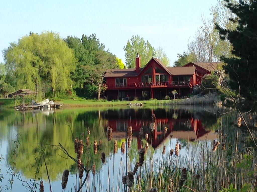 A large red house is reflected on the calm surface of a private lake, surrounded by lush greenery. The property features a spacious deck overlooking the water. Tall reeds and cattails frame the foreground, enhancing the serene natural setting.