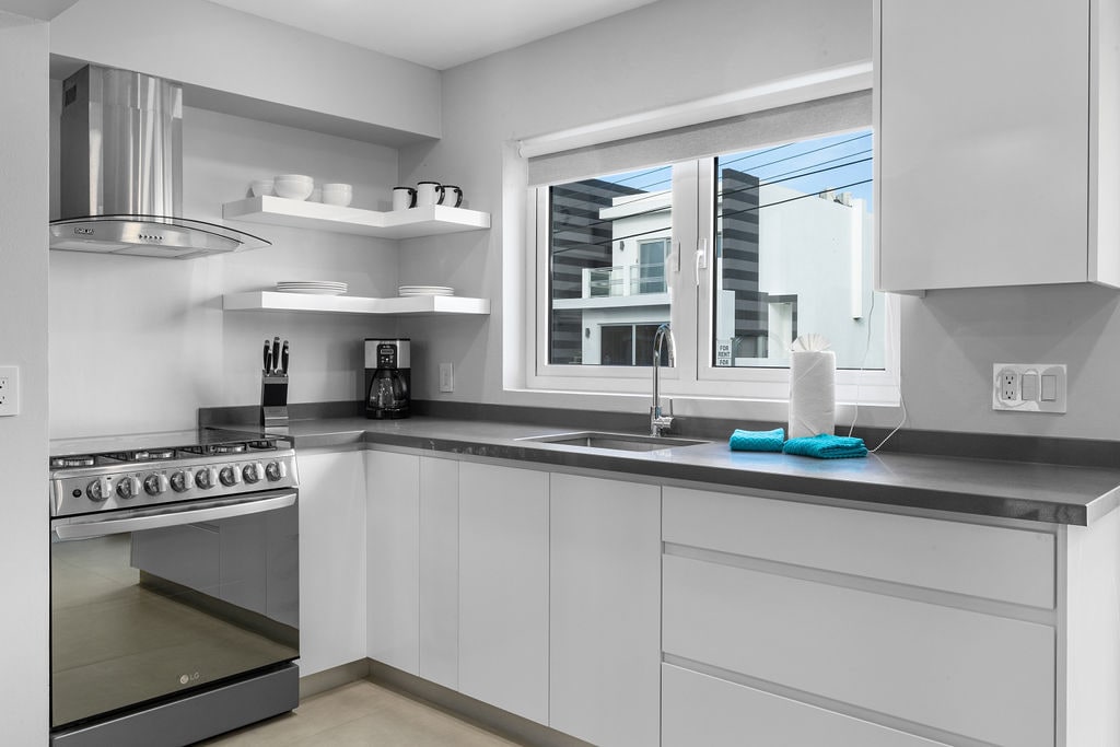 A modern kitchen space features sleek white cabinetry and a gray countertop. Stainless steel appliances, including a gas range and coffee maker, are visible. Natural light streams through a window above the sink, highlighting the minimalist design, while several shelves display dishware.