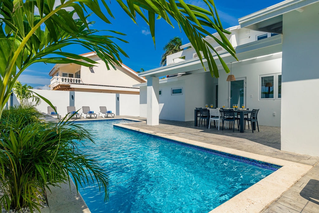 The image showcases a tranquil pool surrounded by palm greenery, with lounge chairs positioned nearby. A covered dining table appears in the foreground, set for outdoor meals. The modern architectural lines of the villa are visible against a clear blue sky.