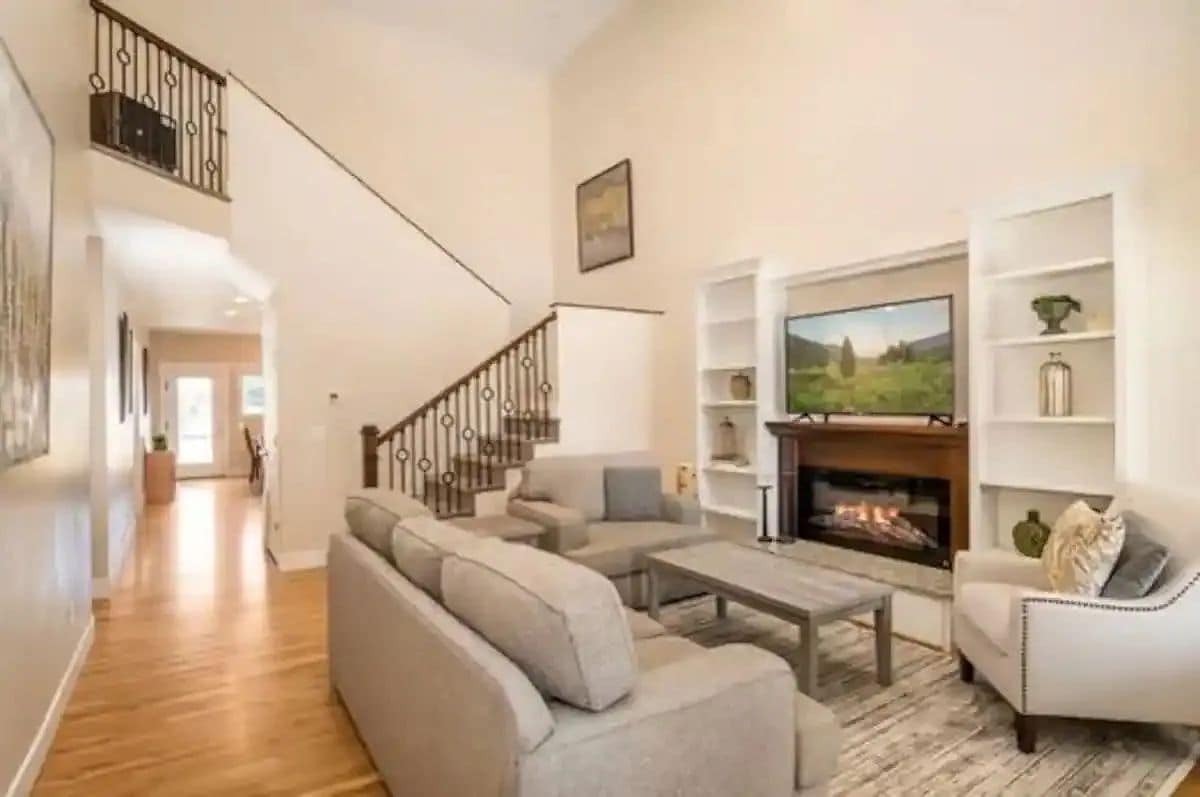 A spacious living area features a cozy gas fireplace beneath a mounted television. Soft gray sofas are arranged around a coffee table atop a light-patterned rug. Elegant shelves showcase decorative items, while a staircase with wrought iron railings leads to the upper level.