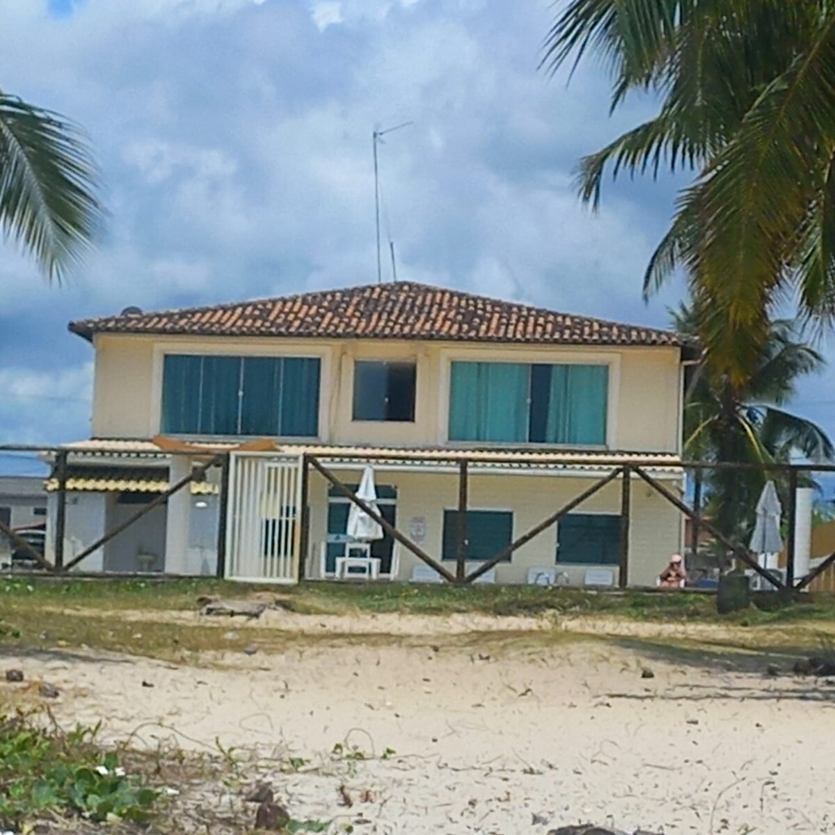 The two-story apartment building features a terracotta tiled roof and multiple windows with blue curtains, set against a sandy beach landscape. Palm trees frame the structure, which is positioned close to the shoreline, creating a relaxed coastal atmosphere.