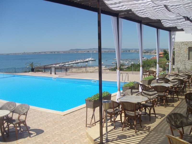 A sparkling pool is situated within the resort grounds, accompanied by a shaded seating area featuring round tables and chairs. The pool area overlooks the bay, offering scenic views of the water and nearby marina, with clear skies enhancing the serene atmosphere.