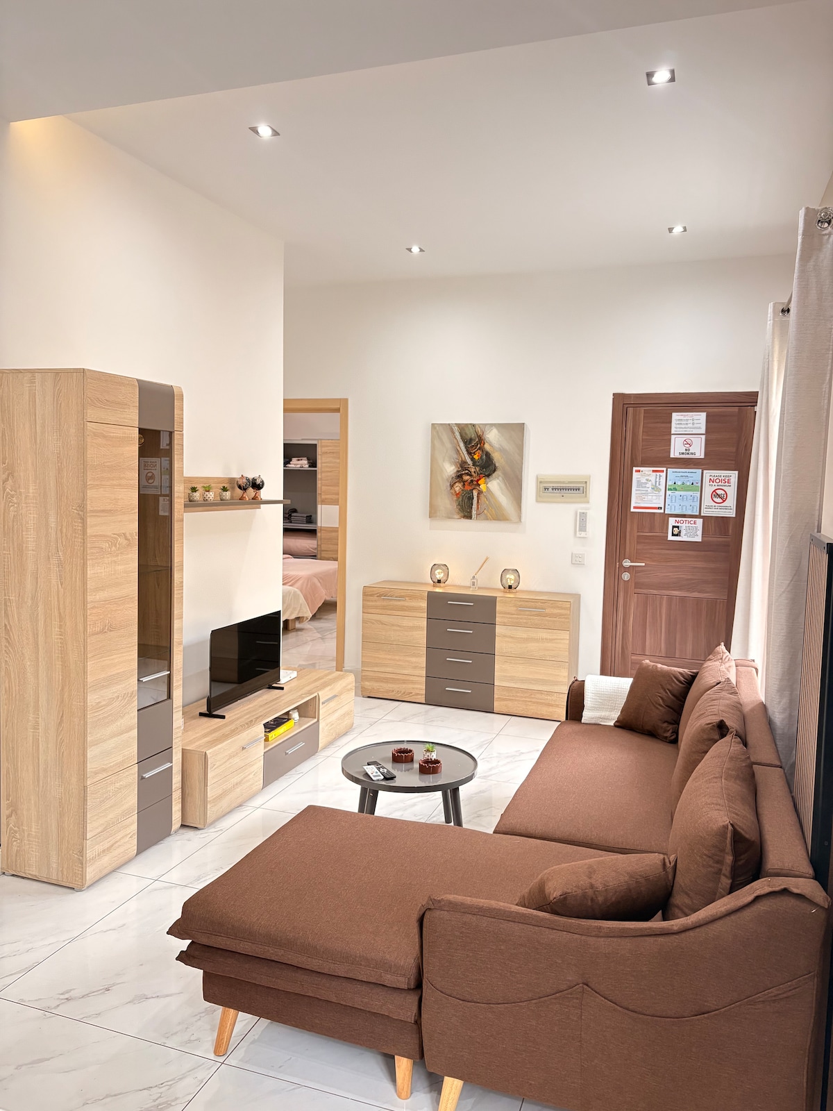 A modern living area is depicted, featuring a brown sofa with wooden legs. A small round table sits in front, complemented by decorative items. Light-colored cabinets and a wall-mounted television enhance the space. Bright overhead lights illuminate the area, creating a welcoming atmosphere.