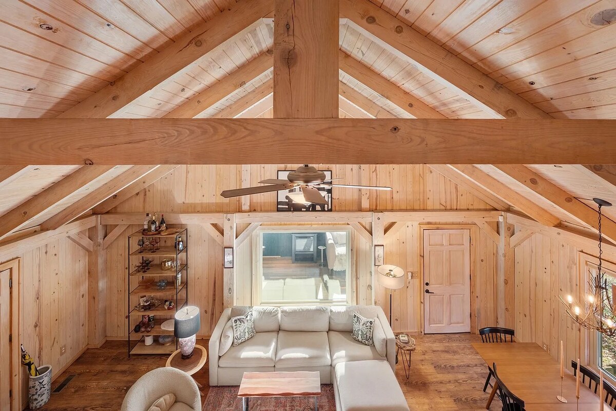 The open-concept living area showcases vaulted wooden ceilings with exposed beams. Natural wood paneling adds warmth to the space. A comfortable sectional sofa is positioned centrally, surrounded by a cozy lounge chair and a dining table set for six, contributing to a welcoming atmosphere.
