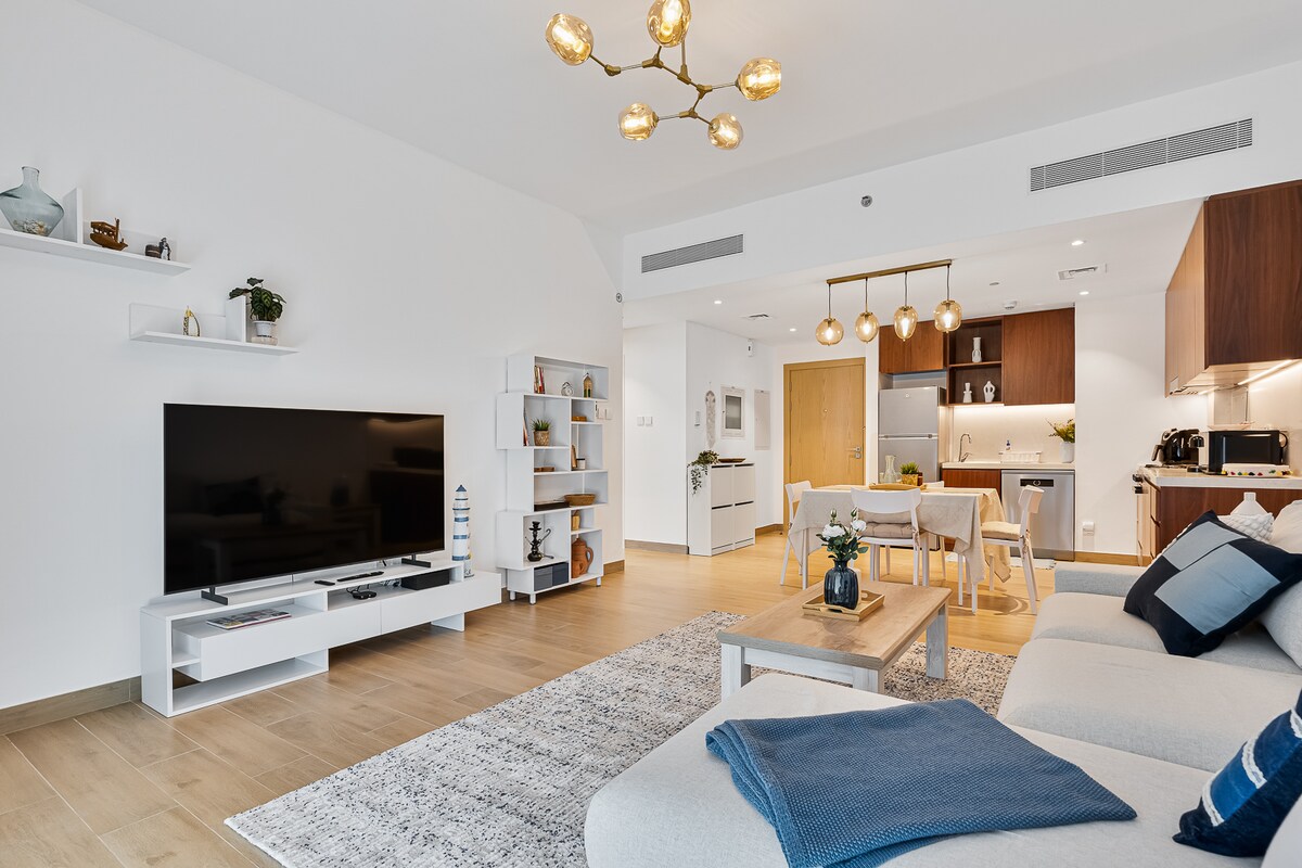 The living room showcases a comfortable seating area with a plush sofa and a contemporary coffee table. A large flat-screen TV is mounted on the wall. The open-concept layout provides a view into the sleek kitchen, accented by warm wooden cabinets and modern lighting fixtures.