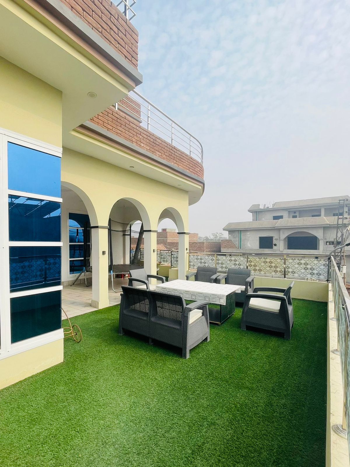 An outdoor terrace is presented with a well-maintained green lawn, featuring a table surrounded by comfortable seating. Arched openings enhance the space, and large windows allow natural light to flow in. A peaceful atmosphere is fostered by the surrounding views of neighboring buildings.