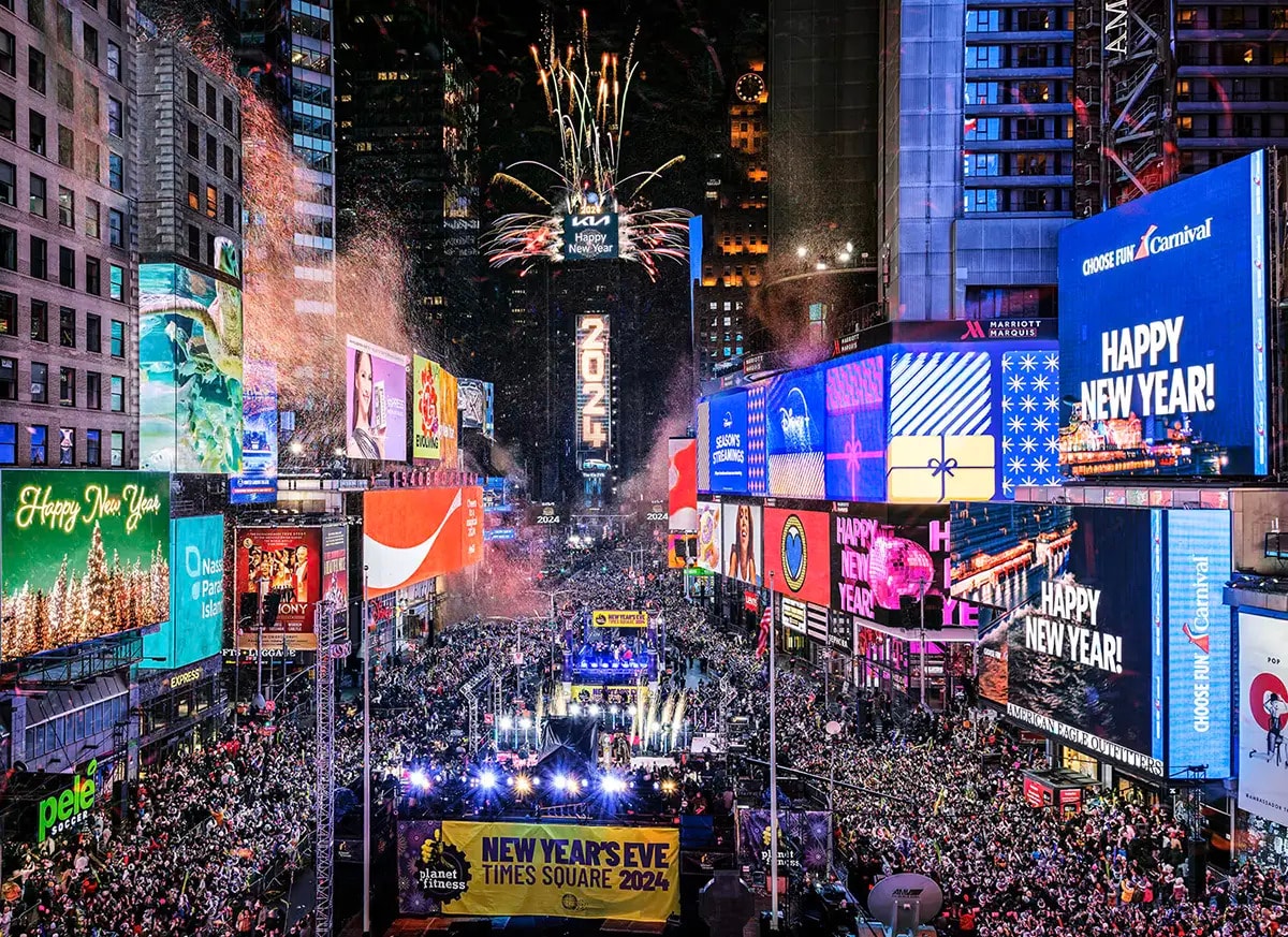 A vibrant New Year's Eve celebration is depicted in Times Square, featuring a large crowd surrounded by bright, illuminated billboards. Colorful fireworks fill the sky above, with various signs displaying festive messages. The scene conveys a lively urban atmosphere filled with energy.