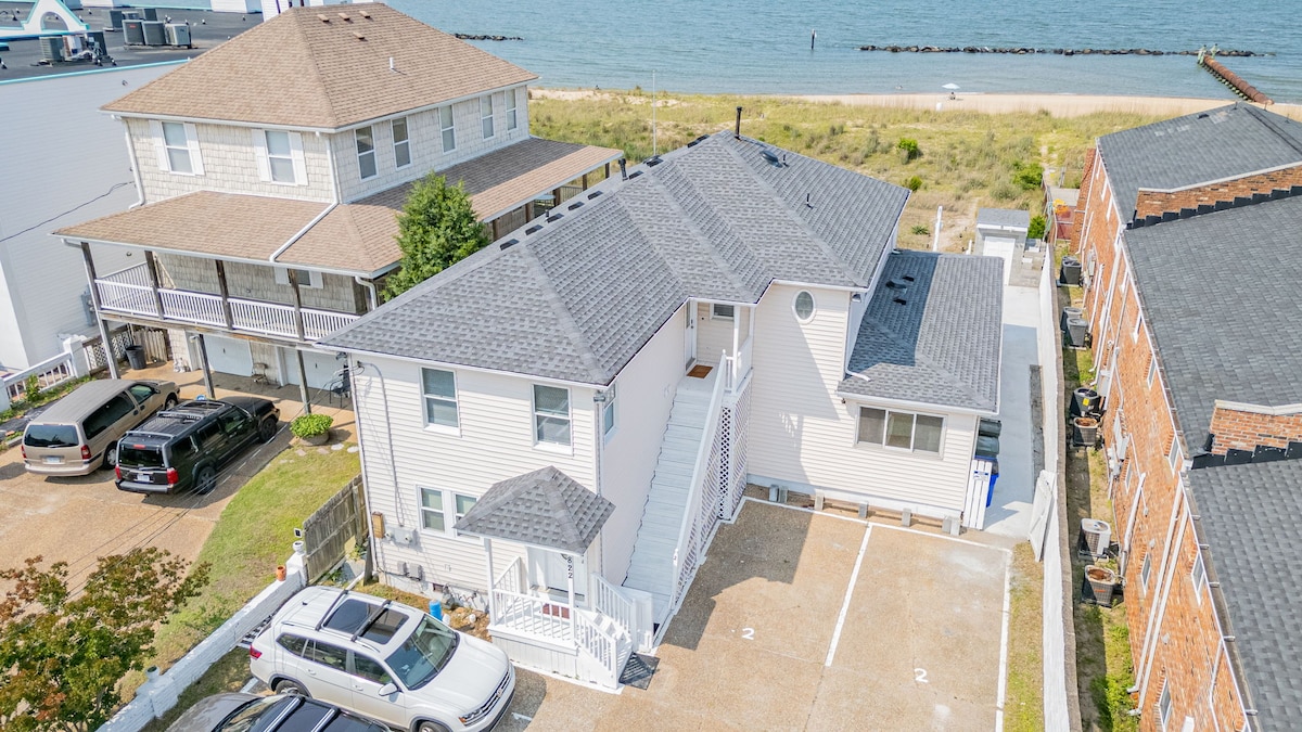 An aerial view captures a beachfront duplex with light-colored siding, surrounded by grass and sand. Parking spaces are visible in front of the units, along with several vehicles. The nearby beach and water are seen in the background, emphasizing direct beach access.