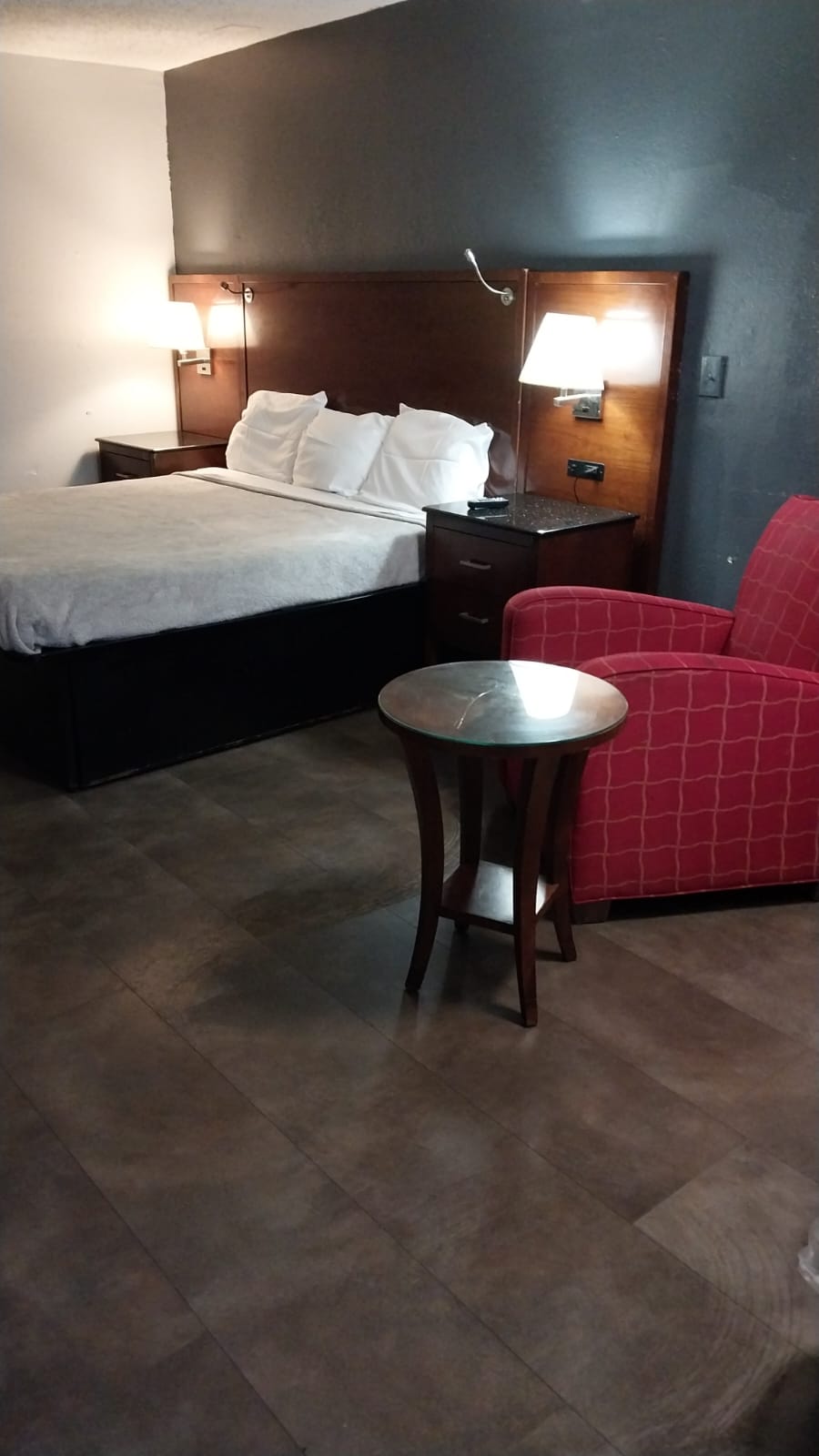 A sleeping area is arranged with a spacious bed adorned with white linens. A nightstand and lamps provide functional lighting. A small side table and a patterned armchair create a cozy seating space. Warm tones in the room enhance the overall comfort.