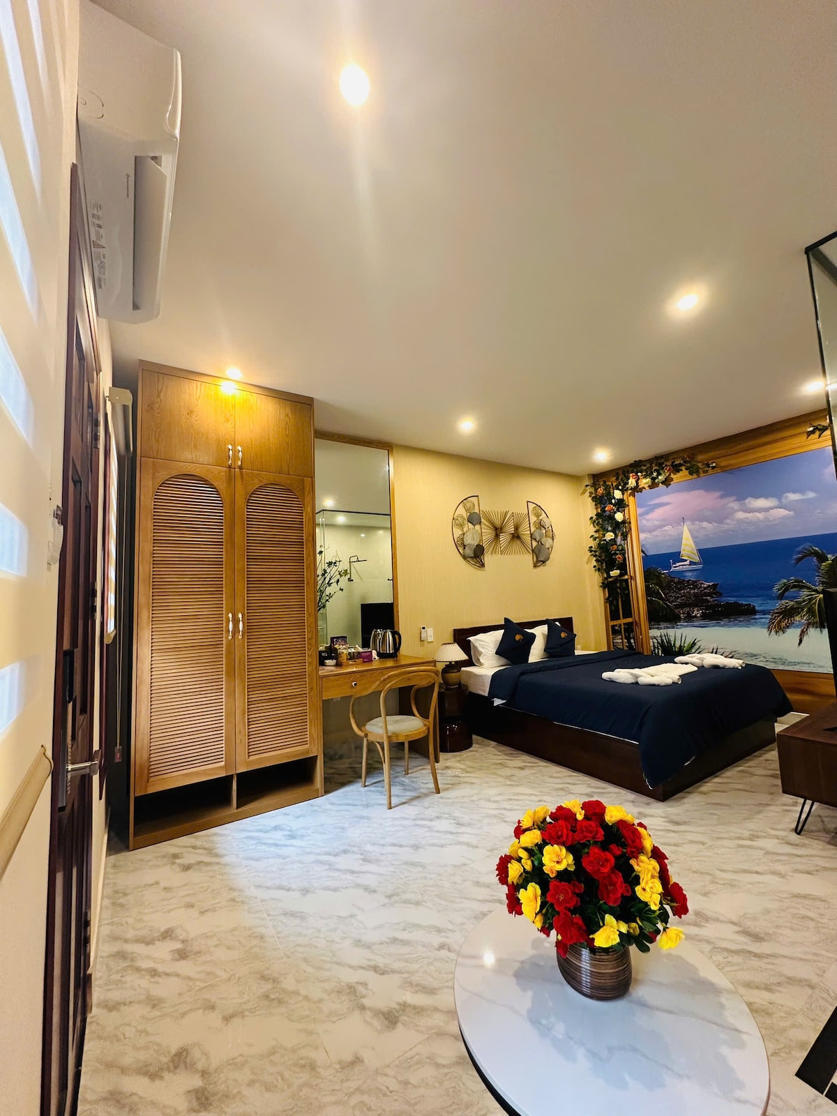 A well-appointed room features a spacious bed with dark linens and an accent mirror framing a scenic mural. A small circular table adorned with a vase of colorful flowers adds a touch of vibrancy, while a wooden wardrobe and makeup area provide functional space.