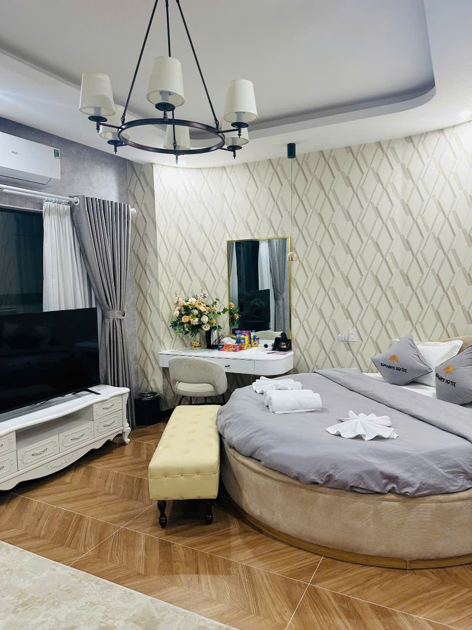 A comfortably appointed bedroom features a round bed covered in soft linens. A mirror reflects a decorative flower arrangement. A television set sits on a stylish cabinet, while a small, light-colored bench offers additional seating options. Natural light enters through window drapes.