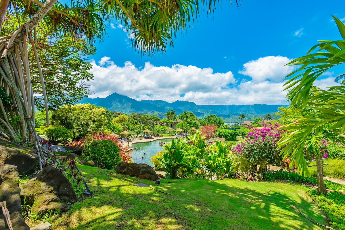 Lush greenery and vibrant tropical flora frame a scenic view of the resort grounds. Majestic mountains rise in the distance, complemented by a tranquil pool area and inviting lounge spaces. The bright blue sky features scattered clouds, creating a serene atmosphere of natural beauty.