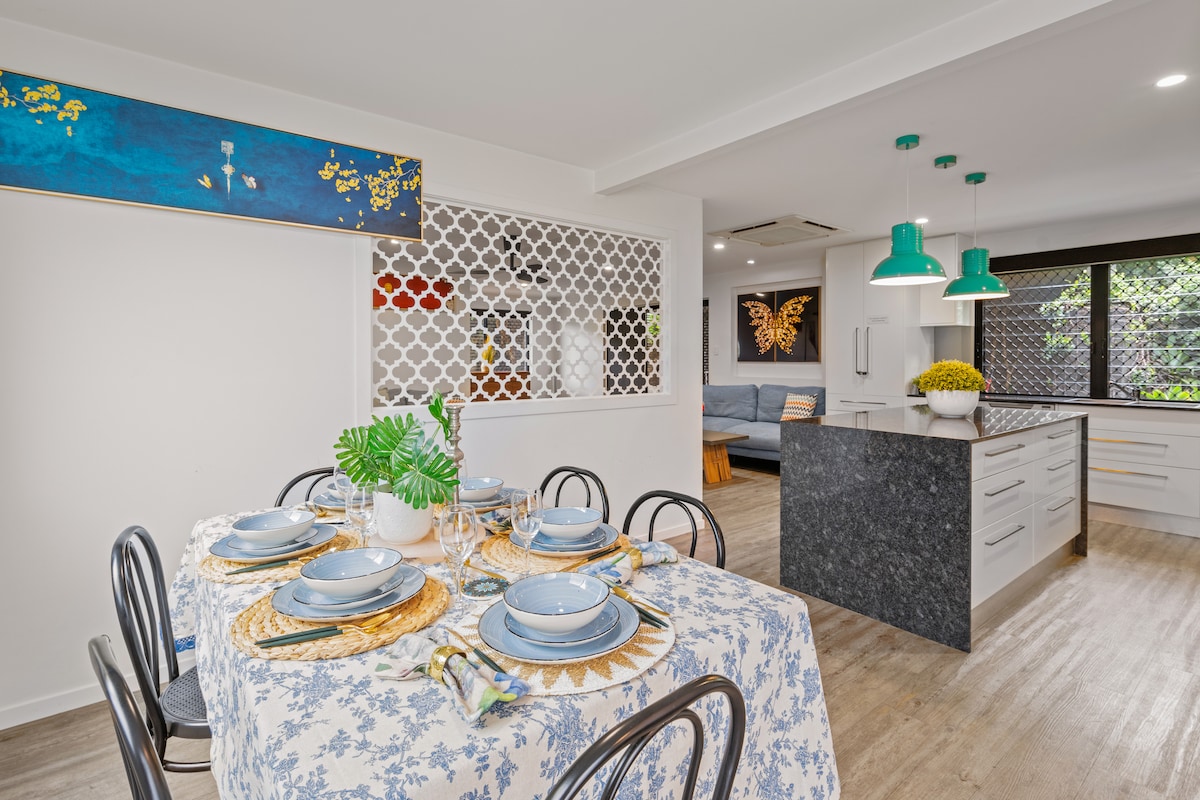 A dining area features a table set with blue dinnerware and decorative accents. A vibrant mural is mounted on the wall, and green pendant lights hang above the table. The open kitchen is visible in the background, showcasing modern cabinetry and appliances.