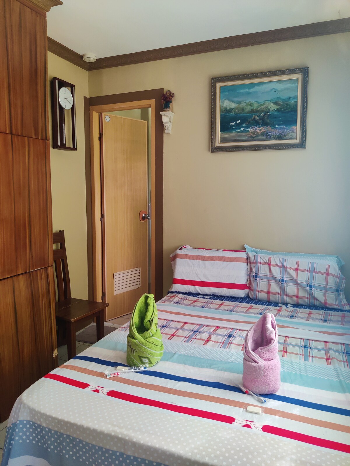 A cozy sleeping area is characterized by a checkered bedding set laid over a spacious bed. Two towel origami figures rest on the bed, adding a playful touch. A wooden chair is positioned against the wall, and a painting is displayed above the bed.