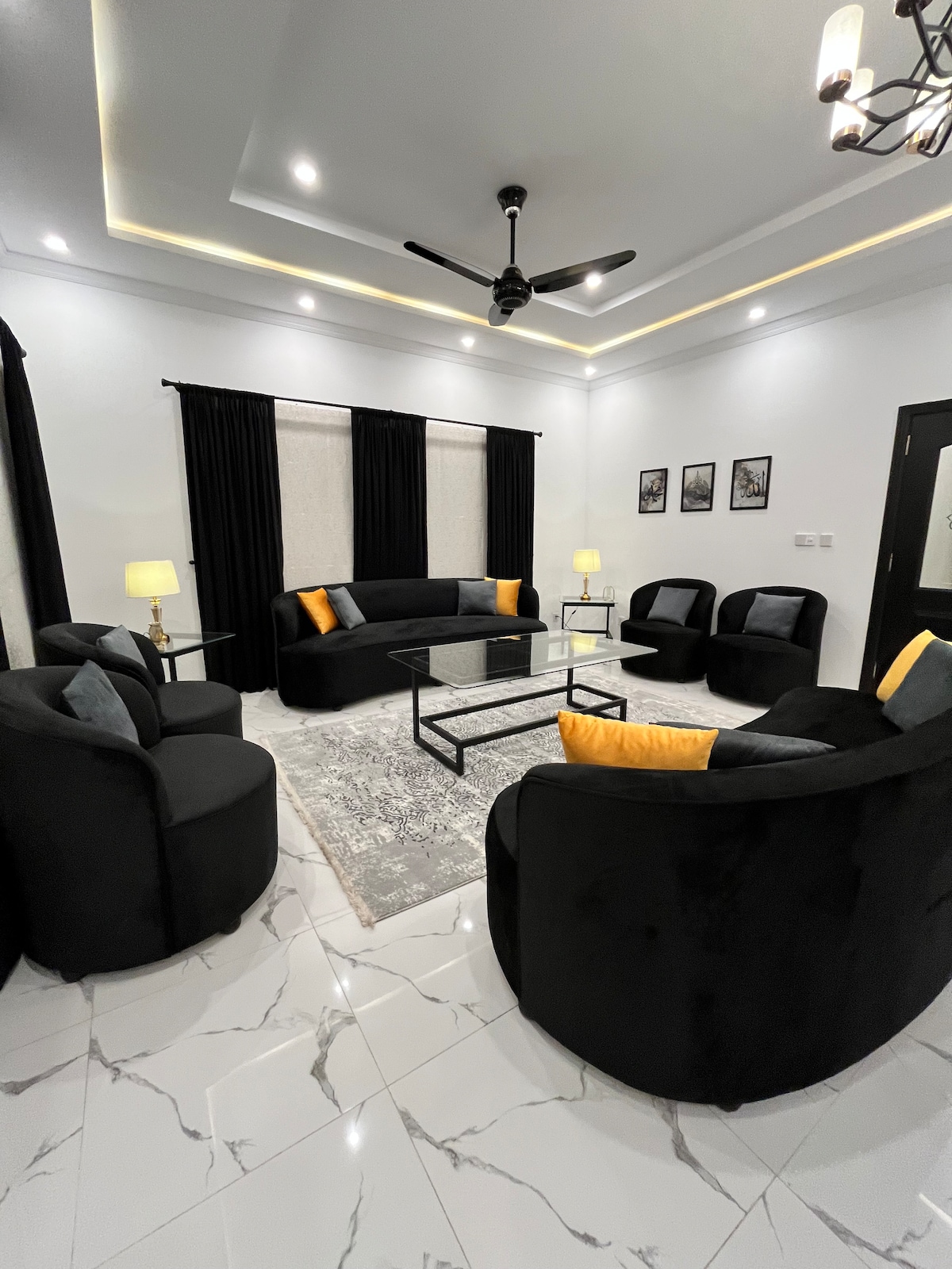 A modern living room is filled with plush black seating, creating a comfortable space for relaxation. Bright yellow accents are present on the cushions. A sleek coffee table sits atop a patterned area rug, while soft lighting from lamps enhances the ambiance.