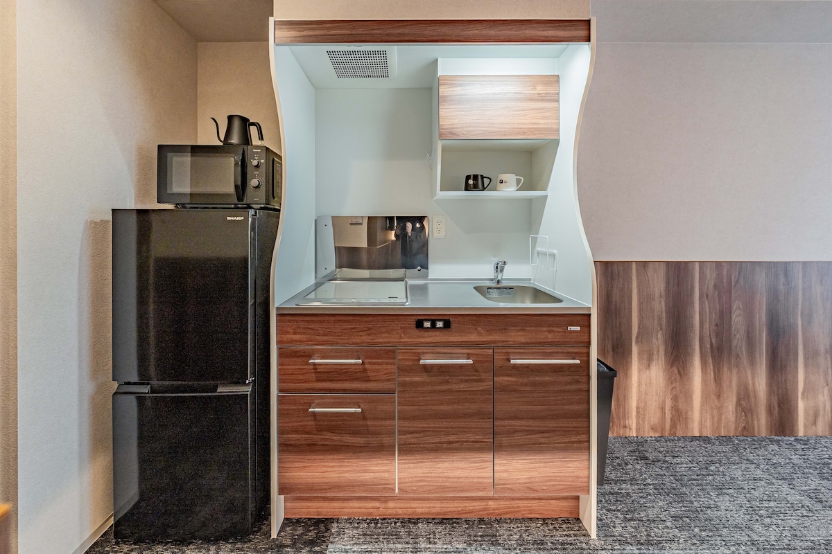 A compact kitchenette is equipped with modern appliances, including a refrigerator, microwave, and sink. Wooden cabinetry provides storage space, while countertop surfaces are available for meal preparation. A coffee mug and utensils are neatly placed on the shelves.