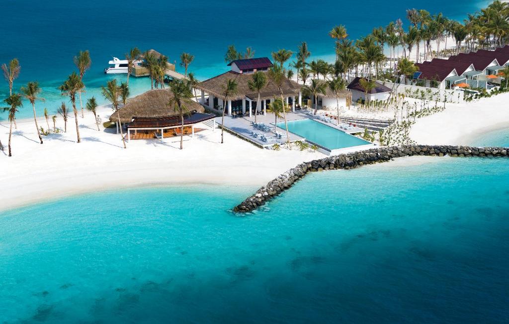 An aerial view of a coastal resort showcases a sandy beach flanked by palm trees and a turquoise lagoon. A swimming pool is centrally located among the resort's buildings, which feature thatched roofs and modern structures, while a pier extends into the water.