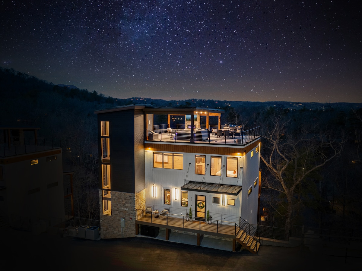 A modern cabin is showcased under a starlit sky, highlighting its spacious rooftop lounge and illuminated exterior. Large windows provide views of the surrounding nature, while multiple outdoor seating areas invite relaxation amidst a serene setting.