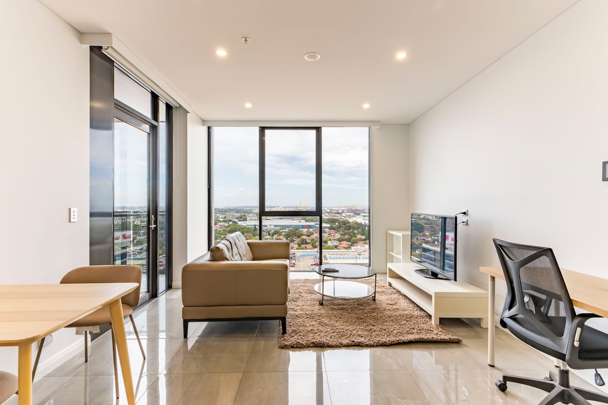 A modern living space is highlighted by floor-to-ceiling windows, offering expansive views of the city below. A comfortable sofa is positioned beside a sleek TV stand, and a round coffee table sits atop a soft area rug. A dining table is visible in the background.
