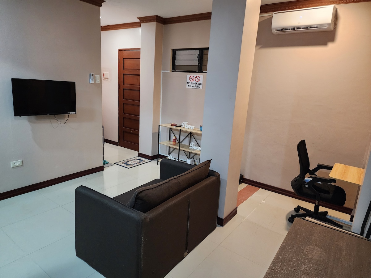The lounge area is furnished with a two-seater sofa, a television mounted on the wall, and an office table with a swivel chair. The space is air-conditioned, and the light-colored floor tiles complement the neutral wall tones. A shelf is positioned against the wall.