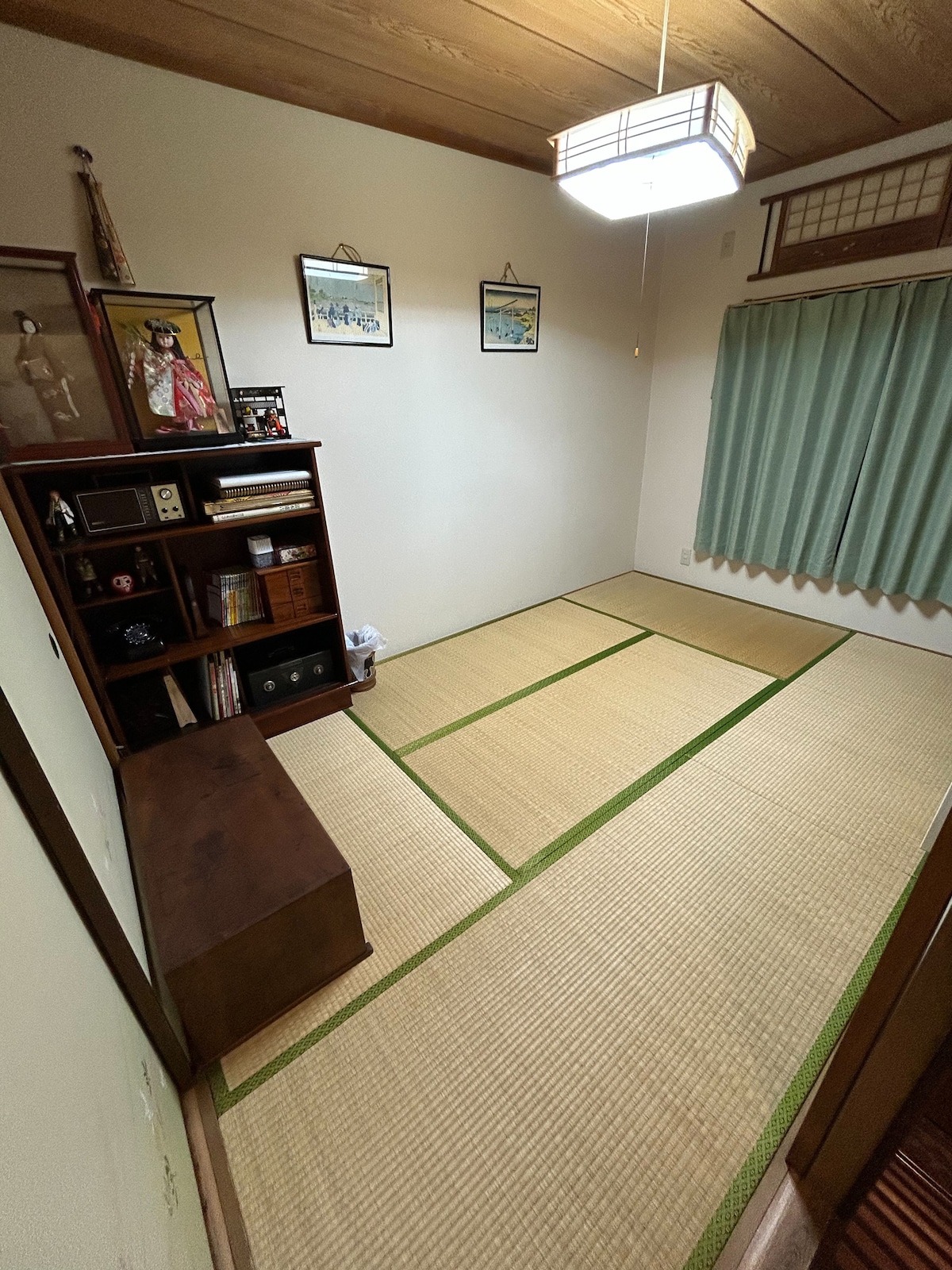A traditional tatami room features wooden accents and soft lighting. A bookshelf with various items is positioned against the wall, while a small bench occupies the corner. Green curtains gently frame the window, providing a serene ambiance in this quiet space.
