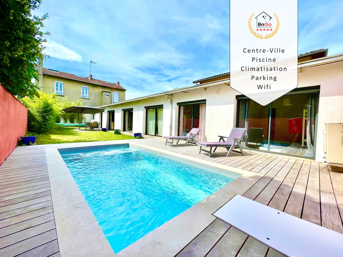 An outdoor area features a private swimming pool set against a backdrop of greenery. Sun loungers are arranged on a wooden deck, while large glass doors provide access to the interior of the villa. A clear sky enhances the inviting ambiance.