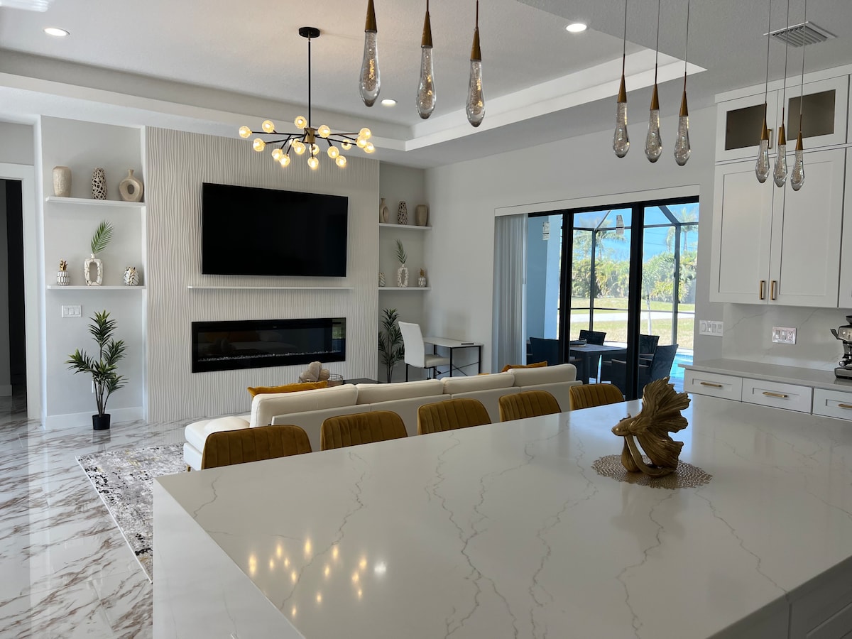 A spacious living area features a large flat-screen TV mounted on a stylish accent wall. Natural light floods the room through expansive glass doors overlooking the outdoors. Elegant pendant lighting and modern decor pieces complement the neutral color palette, enhancing the inviting feel of the space.