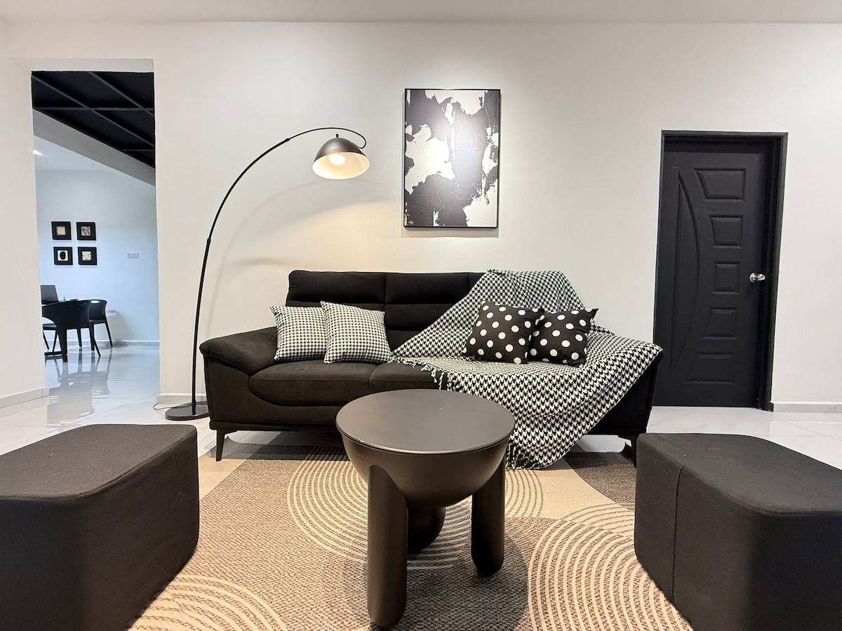 The living area features a modern black sofa adorned with patterned cushions and a cozy throw. A minimalist floor lamp provides illumination, while a round coffee table sits atop a stylish rug. Dark accents are showcased in the wall art and door.
