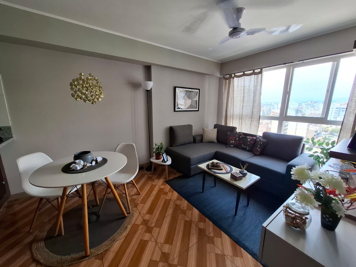 A modern living space features a comfortable sectional sofa adorned with cushions. A small dining table with white chairs is positioned nearby, creating a welcoming area. Natural light enters through large windows, offering a view of the surrounding cityscape. Decorative touches include plants and a stylish light fixture.
