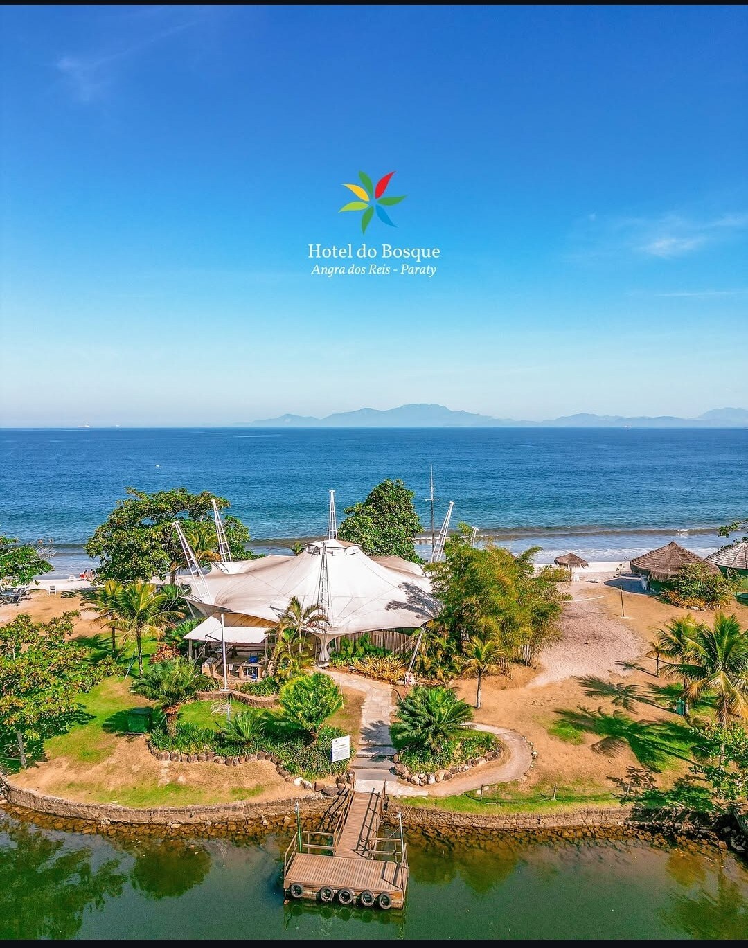 An aerial view of Hotel do Bosque reveals its unique architecture surrounded by lush greenery and sandy beach. The vibrant blue ocean extends in the background, with gentle waves lapping at the shore. A wooden dock leads towards the water, inviting relaxation.