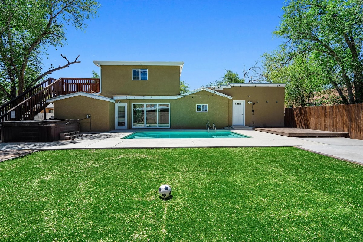 A spacious yard features vibrant artificial turf, complemented by a well-maintained pool and surrounded by a paved area. A two-story structure is visible in the background, with a deck accessible from the upper level and a hot tub situated nearby.