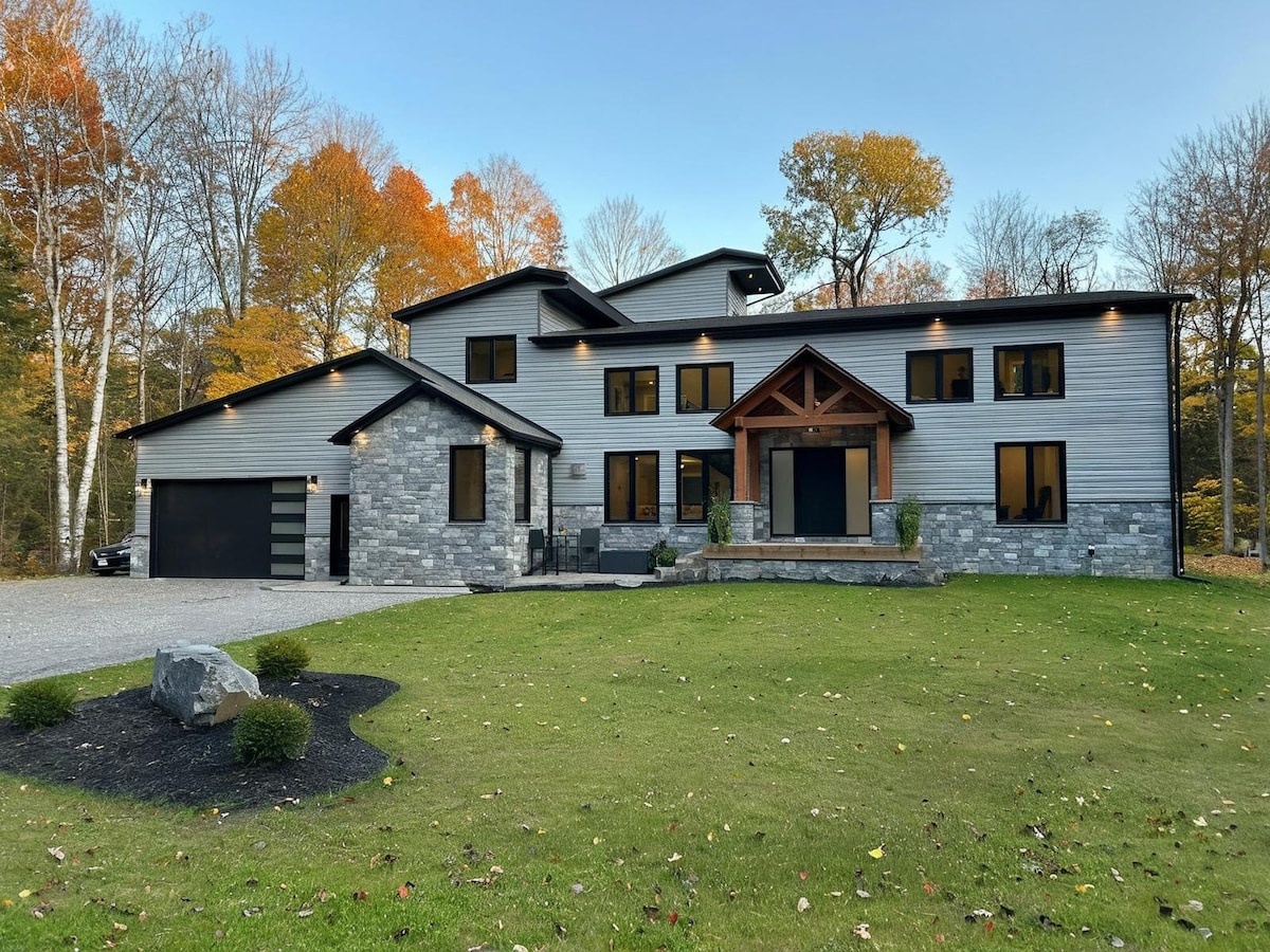 The exterior of a modern mansion is showcased, featuring a combination of stone and metal siding. Large windows provide views into the interior. A front entrance with a wooden overhang and stone steps adds a welcoming touch. Surrounded by trees displaying autumn colors, the manicured lawn is visible.