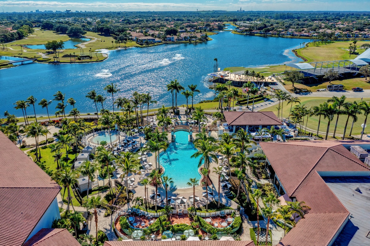Aerial view of a resort-style pool surrounded by palm trees and lounging areas. The clear blue water of the pool contrasts with the lush greenery. A lake and golf course are visible in the background, highlighting the scenic landscape of the PGA National environment.