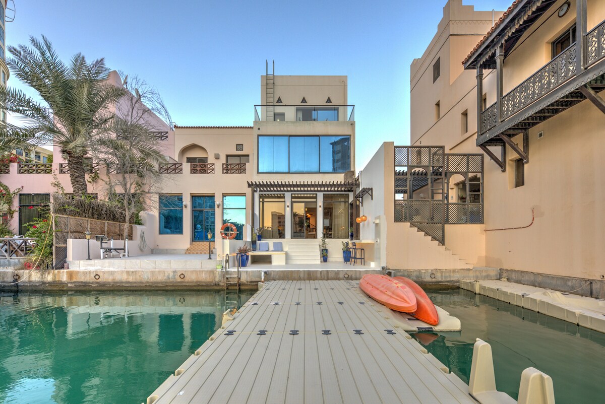 A tranquil waterfront view is captured, featuring a private dock leading to a serene body of water. The modern villa is observed in the background, showcasing large glass windows and a spacious terrace. Two kayaks are positioned at the edge of the dock for easy access.