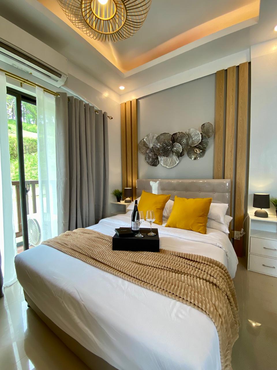 A bright bedroom features a spacious bed layered with soft white linens and a textured beige blanket. Yellow accent pillows are placed at the head of the bed. A decorative wall piece is displayed above, and large windows allow for natural light, framed by sheer curtains.