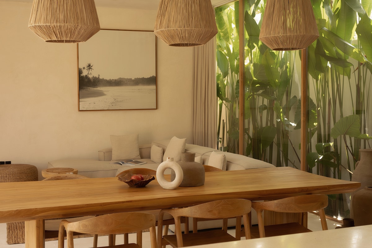 A dining area features a wooden table surrounded by several chairs. Above, three pendant lights are crafted from natural fibers. A large artwork depicting a serene landscape is displayed on the wall. Lush green plants provide a vibrant backdrop, enhancing the indoor space's natural feel.